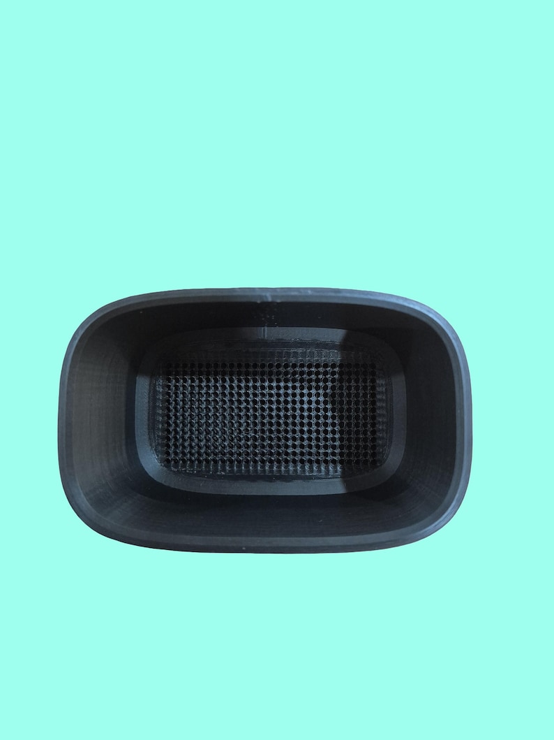 May include: A black, rectangular container with rounded corners and a perforated interior base. The container is set against a light teal background. The design suggests a functional, possibly 3D-printed, item.