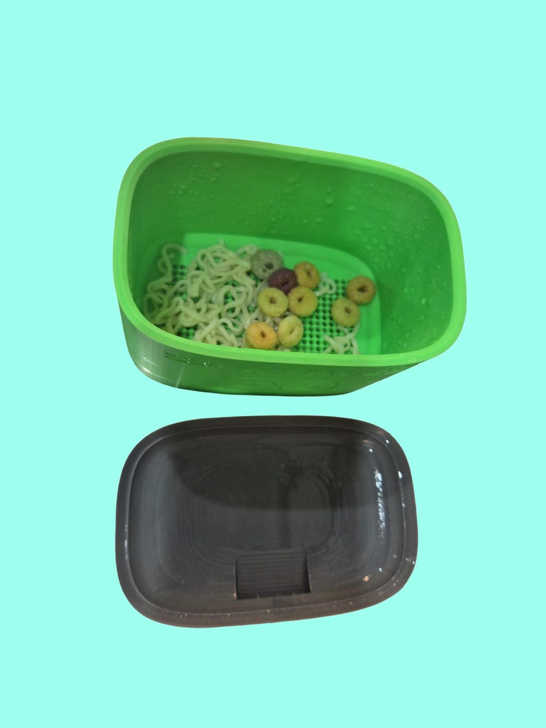 May include: A green plastic container with a dark brown lid. Inside the container are noodles and colorful cereal pieces. The container is on a light blue background.