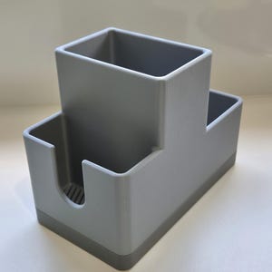 May include: A gray plastic desk organizer with a two-tiered design. The organizer has a rectangular base and a taller, square compartment on top. A smaller compartment and a curved slot are also visible.
