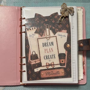 May include: A pink six-ring binder containing a decorative insert with a brown patterned background, a large bow, and the text "DREAM PLAN CREATE" and "Minnette". The insert features illustrations of handbags and a coffee cup, secured by two metal paper clips.