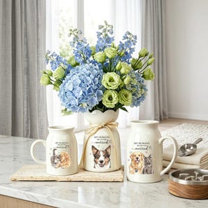 May include: A ceramic vase and two matching mugs featuring illustrations of a dog and cat. The vase holds blue delphiniums and green lisianthus. Each piece displays the text "MY HUMANS ARE GETTING married" with small heart icons and pet portraits.