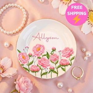 Könnte beinhalten: Eine runde weiße Keramikschale mit goldenem Rand und einem Blumenmuster aus rosa Pfingstrosen mit grünen Blättern. Der Name Allyson ist in rosa Schreibschrift in der Mitte der Schale aufgedruckt. Die Schale ist von verstreuten weißen Perlen umgeben.
