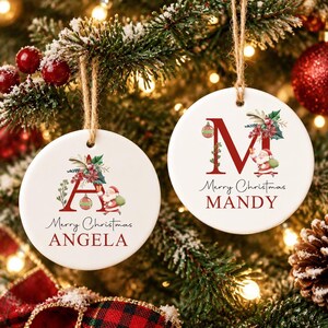 Könnte beinhalten: Zwei runde, weiße Keramikornamente, die an einer Schnur hängen. Das linke Ornament zeigt ein rotes A, eine Weihnachtsmann-Illustration, Stechpalmen und den Text Merry Christmas ANGELA. Das rechte Ornament zeigt ein rotes M und Merry Christmas MANDY.