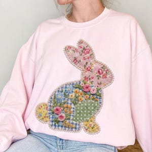 Puede incluir: Una sudadera rosa claro con un diseño de conejo de retazos. El conejo está hecho de varias telas florales, incluyendo rosas rosas, flores azules y amarillas y lunares verdes. El conejo está delineado con un ribete perlado.