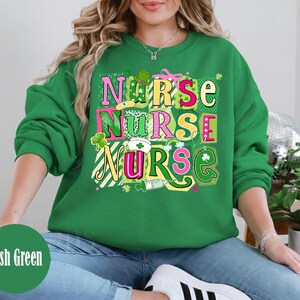 Puede incluir: Sudadera verde con la palabra "NURSE" en letras de bloque coloridas. El diseño incluye tréboles, un gorro de enfermera y una jeringa. La sudadera tiene un tema del Día de San Patricio.