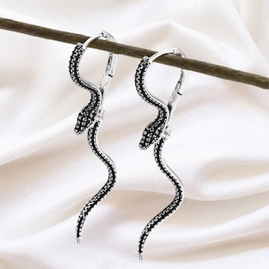 May include: A pair of silver hoop earrings with a snake design. The snakes have black and silver detailing, with small green accents for the eyes. The earrings are displayed against a white backdrop.