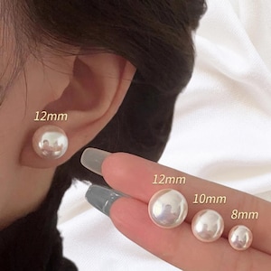 May include: A set of round white pearl stud earrings shown in four sizes. One earring is worn on the earlobe, while three others are held in place, labelled with their respective diameters of 12mm, 12mm, 10mm, and 8mm for size comparison.