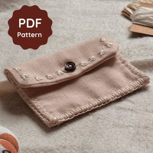 May include: A light pink, handmade fabric wallet with a button closure. The wallet features embroidered floral details and a stitched border. The words "PDF Pattern" are in a brown circle in the upper left corner.