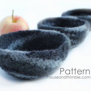 May include: Four gray felted bowls, with a slight blue tint, are arranged in a row. The bowls are made of wool and have a soft, fuzzy texture. The bowls are nested inside each other, with the largest bowl at the back and the smallest bowl at the front. A red apple sits in front of the bowls.