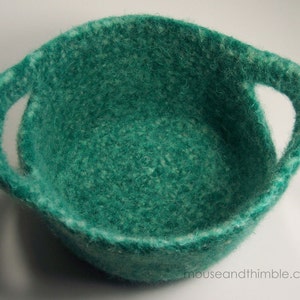 May include: A teal green felted bowl with two handles. The bowl is made of wool and has a soft, fuzzy texture.