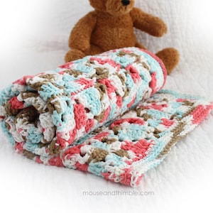 Puede incluir: Una manta de bebé de ganchillo en tonos coral, turquesa, blanco y marrón. La manta está enrollada y tiene un patrón texturizado de cuadrados de la abuela.