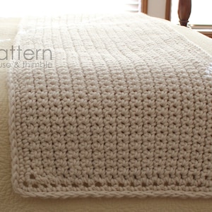 May include: A cream-colored crocheted throw blanket with a simple stitch pattern and a crocheted border. The blanket is lying on a bed.