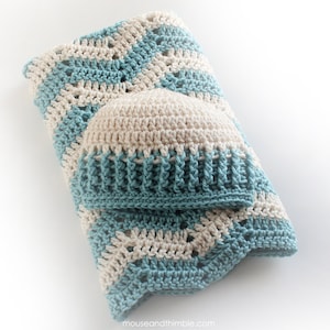 May include: A crocheted baby blanket in blue and cream with a matching crocheted hat. The blanket is folded in half and the hat is sitting on top of it.