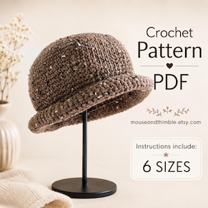 May include: A brown speckled crochet hat displayed on a black stand. The text reads Crochet Pattern PDF, mouseandthimble.etsy.com, and Instructions include: 6 SIZES. The hat features a rounded crown and a short, turned-up brim with textured yarn.