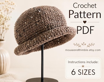 May include: A brown speckled crochet hat displayed on a black stand. The text reads Crochet Pattern PDF, mouseandthimble.etsy.com, and Instructions include: 6 SIZES. The hat features a rounded crown and a short, turned-up brim with textured yarn.