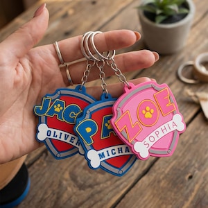 May include: Three personalized shield-shaped keychains in blue, red, and pink. Each keychain features a name, a paw print, and a bone-shaped accent. The keychains are attached to silver key rings.