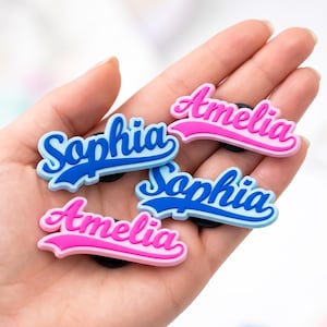 May include: Five colourful shoe charms featuring the names "Sophia" and "Amelia" in a script font. The "Sophia" charms are blue and the "Amelia" charms are pink. Each charm has a black base.