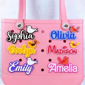 May include: A pink tote bag with decorative name charms. The charms feature names like "Sophia" and "Olivia" in various colors, accompanied by bird-shaped embellishments. The bag has two handles and decorative buttons.