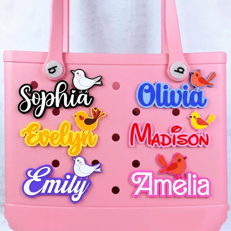May include: A pink tote bag with decorative name charms. The charms feature names like "Sophia" and "Olivia" in various colors, accompanied by bird-shaped embellishments. The bag has two handles and decorative buttons.