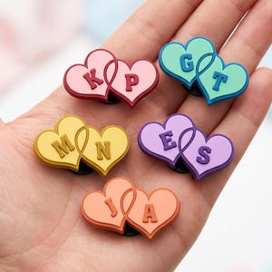 May include: A selection of heart-shaped charms in various colours, each featuring two intertwined hearts with individual initials. The charms are in shades of pink, green, yellow, purple, and orange, with black bases.
