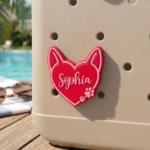 May include: A red and pink heart-shaped charm with the name "Sophia" in cursive script and paw prints. The charm is attached to a beige bag with circular holes. The background includes a pool and sunglasses.