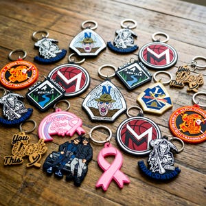 K&ouml;nnte beinhalten: Eine Sammlung von Schl&uuml;sselanh&auml;ngern mit verschiedenen Designs, darunter ein rosa Band, ein Basketball mit dem Buchstaben "M" und milit&auml;rische Embleme. Die Schl&uuml;sselanh&auml;nger sind aus Gummi und Metall gefertigt, mit einer Vielzahl von Farben und Texten.