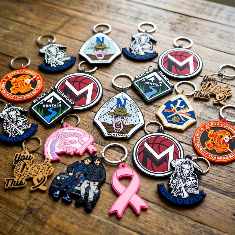 K&ouml;nnte beinhalten: Eine Sammlung von Schl&uuml;sselanh&auml;ngern mit verschiedenen Designs, darunter ein rosa Band, ein Basketball mit dem Buchstaben "M" und milit&auml;rische Embleme. Die Schl&uuml;sselanh&auml;nger sind aus Gummi und Metall gefertigt, mit einer Vielzahl von Farben und Texten.