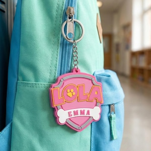 May include: A pink and yellow shield-shaped keychain with the name "LOLA" and "EMMA" printed on it. The keychain is attached to a silver zipper pull on a teal and blue backpack. The keychain has a paw print and bone design.