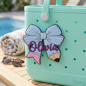 Puede incluir: Un bolso tote de color verde azulado claro con una etiqueta con el nombre en forma de lazo que dice "Olivia". El lazo es azul claro con rayas blancas y detalles en rosa. Un dise&ntilde;o de l&aacute;piz est&aacute; en el lado derecho del lazo. Gafas de sol y una toalla enrollada en el fondo.