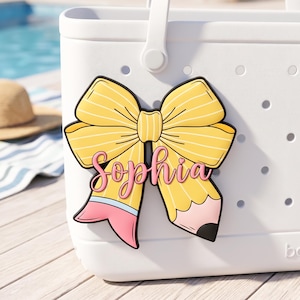 Puede incluir: Un bolso tote blanco con un charm en forma de lazo amarillo. El charm presenta un dise&ntilde;o de l&aacute;piz con el nombre "Sophia" en escritura rosa. El lazo tiene rayas amarillas y contornos negros. El bolso tiene un asa blanca y un patr&oacute;n de puntos.