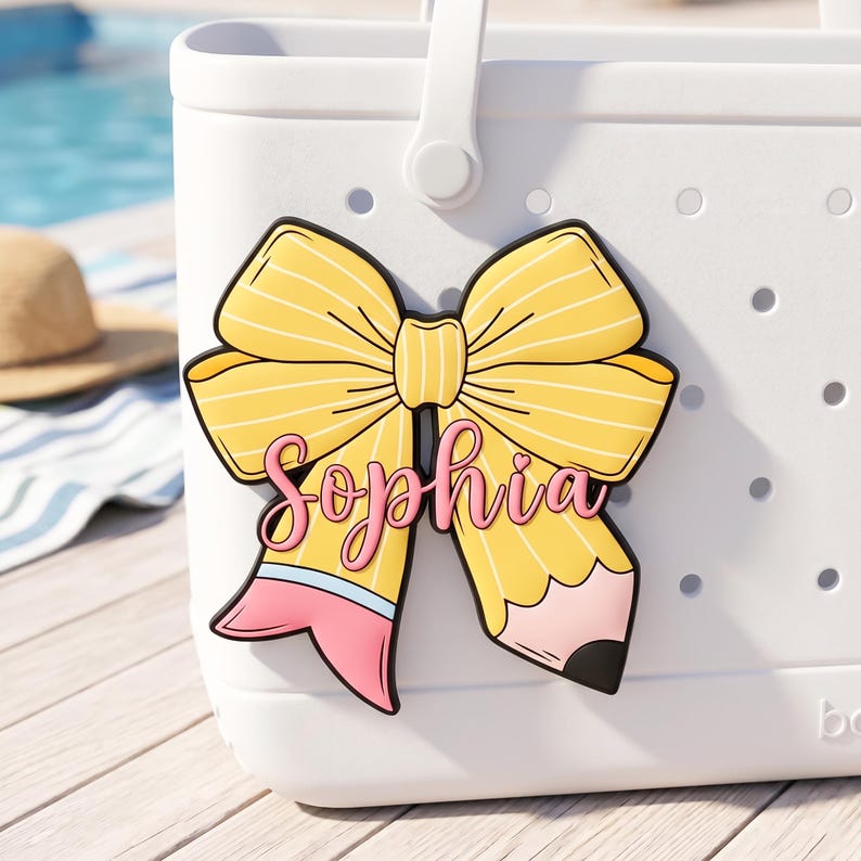 Puede incluir: Un bolso tote blanco con un charm en forma de lazo amarillo. El charm presenta un dise&ntilde;o de l&aacute;piz con el nombre "Sophia" en escritura rosa. El lazo tiene rayas amarillas y contornos negros. El bolso tiene un asa blanca y un patr&oacute;n de puntos.