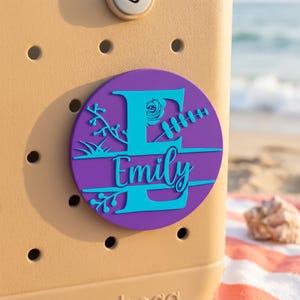 Könnte beinhalten: Ein personalisiertes rundes Emblem in Lila und Türkis mit dem Namen "Emily" und floralen Akzenten. Das Emblem ist an einer beigefarbenen Tasche mit kreisförmigen Löchern befestigt. Der Hintergrund zeigt eine Strandszene mit Meer und Sand.