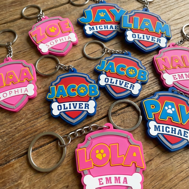 May include: A collection of colorful, personalized keychains. Each keychain features a shield design with a name and a bone-shaped banner. The keychains are in shades of pink, blue, and red, with silver key rings.
