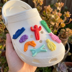 May include: A white foam clog shoe featuring a collection of colorful, stylized human-shaped charms in various poses. The charms are in shades of blue, purple, orange, red, green, pink, yellow, and light blue, attached to the shoe's ventilation holes.