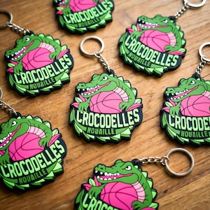 K&ouml;nnte beinhalten: Schl&uuml;sselanh&auml;nger mit einem gr&uuml;nen Alligator &uuml;ber einem rosa Basketball, mit dem Text "CROCODELLES NOUAILLE" in Wei&szlig;. Die Schl&uuml;sselanh&auml;nger haben einen schwarzen Rand und silberne Ringe. Das Design wiederholt sich.
