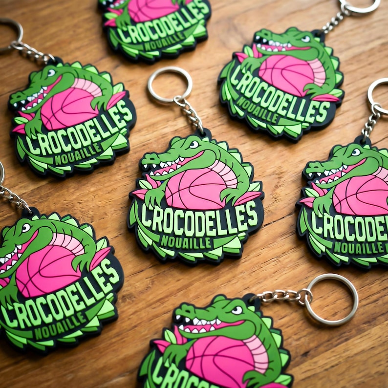 K&ouml;nnte beinhalten: Schl&uuml;sselanh&auml;nger mit einem gr&uuml;nen Alligator &uuml;ber einem rosa Basketball, mit dem Text "CROCODELLES NOUAILLE" in Wei&szlig;. Die Schl&uuml;sselanh&auml;nger haben einen schwarzen Rand und silberne Ringe. Das Design wiederholt sich.