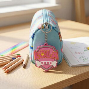 May include: A light blue pencil case with a zipper, featuring a pattern of cartoon characters. A pink shield-shaped keychain with the name "LOLA" and "EMMA" hangs from the zipper. Colored pencils and a notebook are nearby.