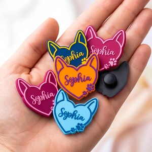 May include: A collection of colorful shoe charms in the shape of a cat's head and heart. Each charm features the name "Sophia" and paw prints. Colors include blue, orange, pink, and black. These charms are designed to be attached to shoes.