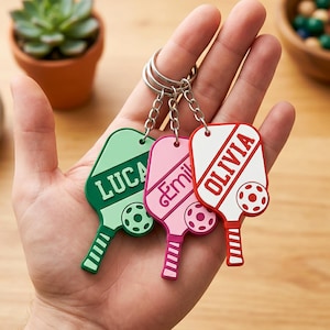 Puede incluir: Tres llaveros personalizados con forma de paleta de pickleball en verde, rosa y blanco. Cada llavero presenta un nombre: "LUCA", "Emil" y "OLIVIA", junto con un diseño de pelota de pickleball. Los llaveros están unidos a llaveros plateados.