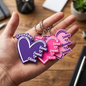 Op de afbeelding: Een hand toont vier hartvormige sleutelhangers in paars en roze. Elke sleutelhanger heeft een witte rand en bevat namen zoals "Emma" en "Lucas". De sleutelhangers zijn bevestigd aan zilveren ringen.