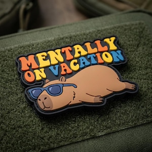 May include: A rectangular morale patch featuring a brown capybara wearing blue sunglasses, lying down. Above the capybara, the text MENTALLY ON VACATION is displayed in bold, multi-colored letters including orange, yellow, and blue against a black background.