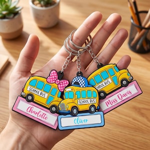 Op de afbeelding: Drie schoolbus-sleutelhangers met metalen ringen. Eén bus heeft een roze strik, één een blauwe pet en één is effen. Elke bus is voorzien van de tekst 'SCHOOL BUS' en een rechthoekig naamplaatje met de namen Charlotte, Oliver en Miss Davis.
