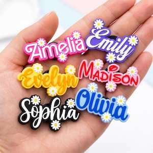 Può includere: Una collezione di ciondoli con nomi colorati, ognuno con un nome diverso in un carattere giocoso. I nomi includono Amelia, Emily, Evelyn, Madison, Sophia e Olivia. Ogni ciondolo è adornato con piccoli dettagli a margherita.