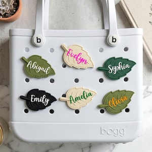Può includere: Una borsa tote bianca con targhette a forma di foglia. Le targhette sono in vari colori, tra cui verde, nero e crema, ognuna con un nome diverso in corsivo. La borsa ha manici bianchi e il marchio "bogg" è visibile.