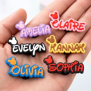 Puede incluir: Una colección de coloridos dijes con nombres, cada uno con un nombre diferente en una fuente lúdica. Los dijes incluyen nombres como Amelia, Claire, Evelyn, Hannah, Olivia y Sophia, con un diseño inspirado en Mickey Mouse.
