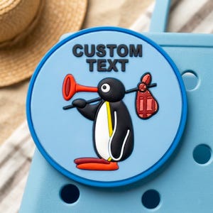 Puede incluir: Un dije redondo azul claro con un diseño de pingüino de dibujos animados. El pingüino es negro, blanco y amarillo, sosteniendo una bocina roja y un saco rojo. El texto "CUSTOM TEXT" está impreso en la parte superior. El dije está sobre un fondo azul claro.