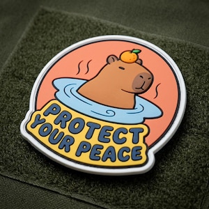 May include: A circular morale patch featuring a brown capybara with a small orange on its head, soaking in a light blue pool against an orange background. The text PROTECT YOUR PEACE is printed in dark blue letters on a yellow banner at the bottom.