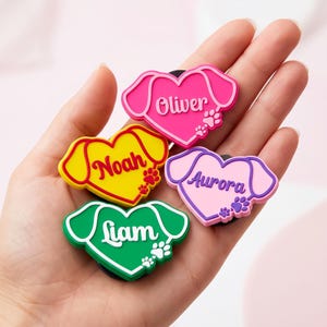 May include: Four colourful dog-shaped charms with names: Oliver, Noah, Aurora, and Liam. Each charm has a heart-shaped centre with a dog's head outline and paw prints. The charms are in pink, yellow, purple, and green.
