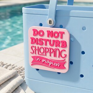 Könnte beinhalten: Hellblaue Tragetasche mit einem rosa rechteckigen Anhänger mit der Aufschrift "DO NOT DISTURB SHOPPING in progress" in fetter, rosa Schrift. Die Tasche hat einen Griff und einen runden Knopf mit dem Buchstaben "B".