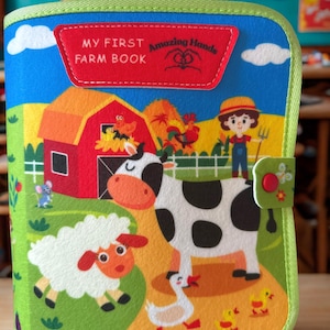 May include: A colorful fabric book titled "MY FIRST FARM BOOK" with illustrations of farm animals and scenes. The cover features a red barn, a cow, a sheep, and a farmer. The book is trimmed with green binding.